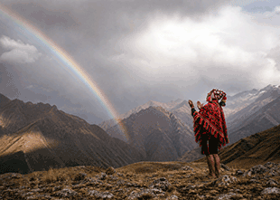 Hiker in a red poncho on a rocky ridge, pointing toward a rainbow over snowy mountains.
