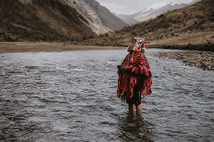 Person wrapped in a red patterned poncho standing knee-deep in a river with mountains in the background.