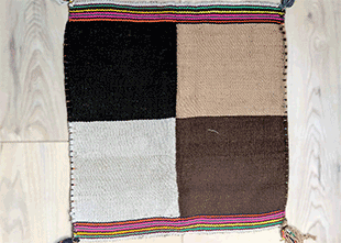 Uncuna | green tara college Small handwoven patchwork rug on a light wood floor with four color blocks: black, beige, gray, and brown, and multicolor striped borders.